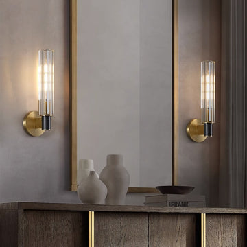A modern room showcases a minimalist design with two Brass Glass Bedroom Lamps flanking a large gold-framed mirror. Below, an E14 LED warmly illuminates the wooden console table adorned with two ceramic vases and a shallow dish.