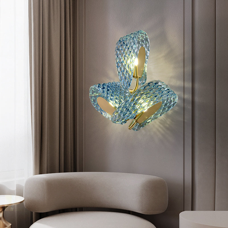 A modern artistic wall lamp featuring wavy blue glass and a golden metal bracket, with a sculptural and elegant design. The light diffuses soft shadows through the glass. Installed on a light-colored wall, the lamp is accompanied by a light-toned curved sofa and a small round metal side table, creating a stylish and minimalist aesthetic perfect for decorating living rooms or lounge areas.