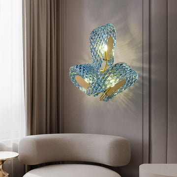 A modern artistic wall lamp featuring wavy blue glass and a golden metal bracket, with a sculptural and elegant design. The light diffuses soft shadows through the glass. Installed on a light-colored wall, the lamp is accompanied by a light-toned curved sofa and a small round metal side table, creating a stylish and minimalist aesthetic perfect for decorating living rooms or lounge areas.