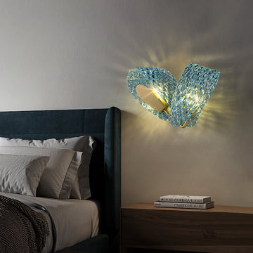 A modern wall lamp installed in a bedroom, featuring wavy blue glass and a golden metal bracket with an elegant design. The light emits soft shadows through the glass, creating a warm and cozy ambiance. Adjacent to the lamp is a bed with a deep green headboard and a wooden bedside table, making it a perfect addition for bedroom decoration and lighting.