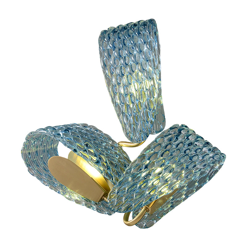 A modern artistic wall lamp featuring wavy blue glass and a golden metal bracket, with a sculptural and elegant design. The light diffuses soft shadows through the glass. Installed on a light-colored wall, the lamp is accompanied by a light-toned curved sofa and a small round metal side table, creating a stylish and minimalist aesthetic perfect for decorating living rooms or lounge areas.