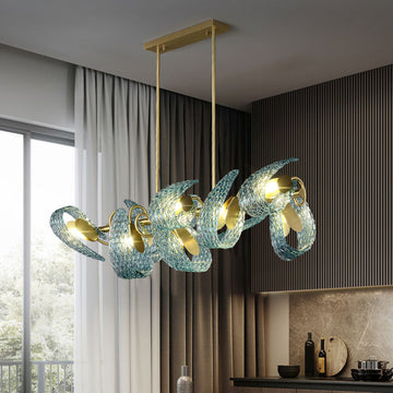 A modern dining room features a unique chandelier with twisting blue glass designs and gold accents. The room has a large window with sheer curtains, a striped wall, and a countertop with various items. Natural light illuminates the space.