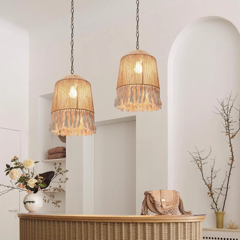 The Bohemian Tassel Chandelier features three beige cylindrical pendant lights with white ruffled edges and textured shades. Suspended by thin chains, it emits a warm glow against a neutral backdrop, enhancing the Bohemian aesthetic.