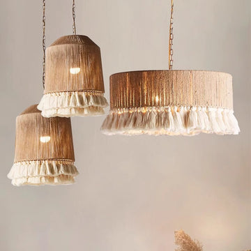 The Bohemian Tassel Chandelier features three beige cylindrical pendant lights with white ruffled edges and textured shades. Suspended by thin chains, it emits a warm glow against a neutral backdrop, enhancing the Bohemian aesthetic.