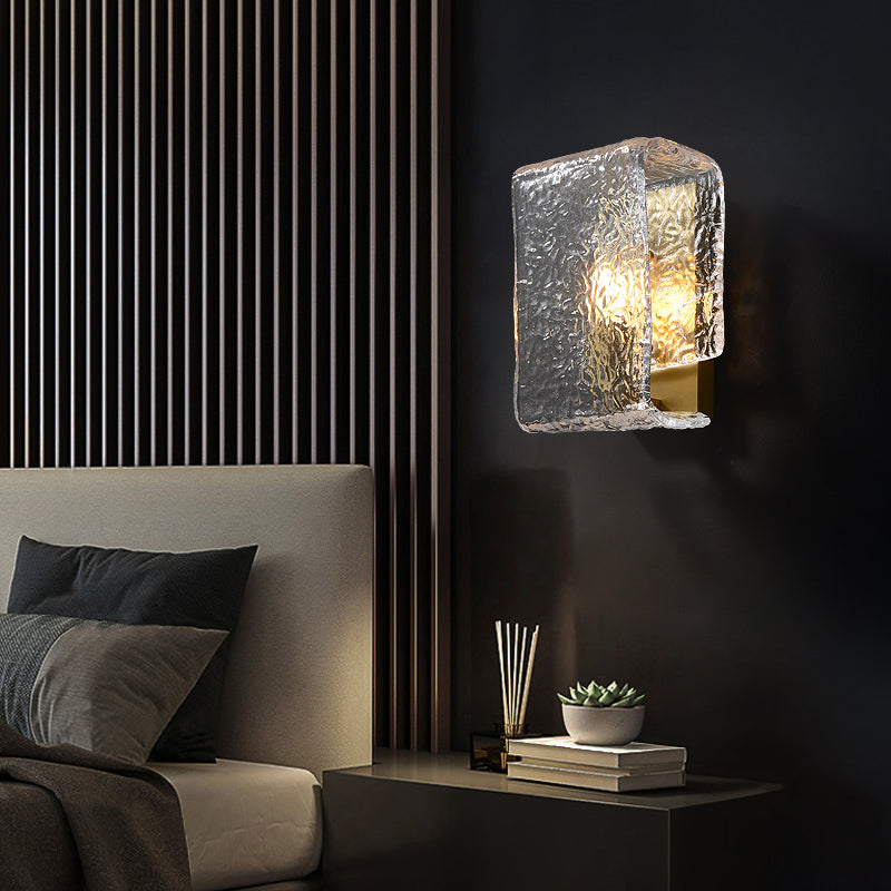 Modern textured wall sconce illuminates beside a bed with gray bedding and small bedside decor.