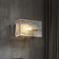 Textured glass wall sconce illuminated against a gray wall with dark curtains to the side.