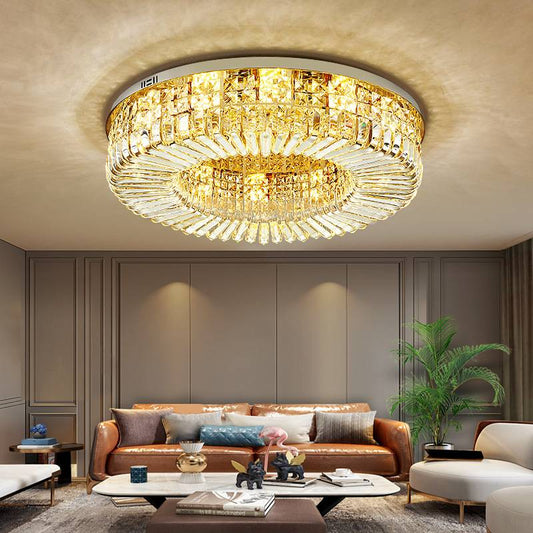 Elegant living room showcasing a large, ornate Ceiling Round Crystal Lamp with gold accents. The space includes a brown leather sofa, modern coffee table adorned with decor items, and a green plant in the corner, all set against beige and gray-toned walls.