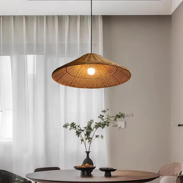 A cozy dining area showcases a round table with a black vase of greenery and a sleek black bowl. Above, a Cuban rattan chandelier emits a warm glow with its LED bulb. Sheer white curtains cover large windows, softly diffusing natural light.