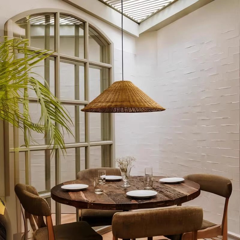 A cozy dining area showcases a round table with a black vase of greenery and a sleek black bowl. Above, a Cuban rattan chandelier emits a warm glow with its LED bulb. Sheer white curtains cover large windows, softly diffusing natural light.