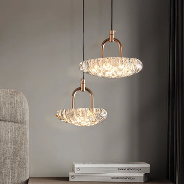 Two elegant Ice pattern chandeliers with textured glass shades hang from the ceiling on adjustable wires, emitting a warm glow. They feature gold-colored fittings, while below them, gray textured fabric is partially visible next to a stack of two books.
