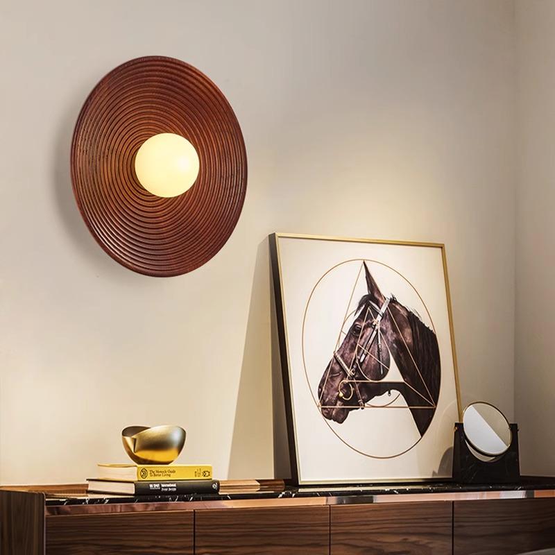 The Japanese Mahogany Wall Lamp, circular and featuring a G9 LED bulb, is mounted on a light wall. Nearby, sheer curtains let soft light through a window. A beds headboard with woven rattan design is partially seen.
