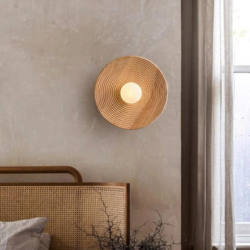 The Japanese Mahogany Wall Lamp, circular and featuring a G9 LED bulb, is mounted on a light wall. Nearby, sheer curtains let soft light through a window. A beds headboard with woven rattan design is partially seen.