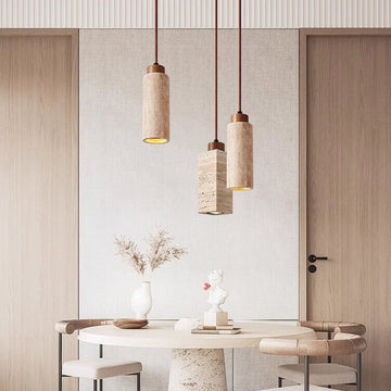 A minimalist dining area showcases a round table blending travertine and wood with beige walls. Above, three Travertine Restaurant Chandeliers provide dimmable LED lighting. A white vase of dried branches and a small sculpture adorn the table, flanked symmetrically by two wooden doors.