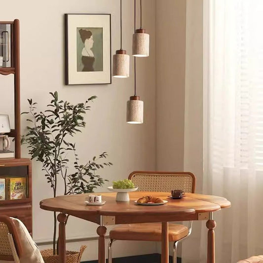 A cozy dining area showcases a round wooden table with woven chairs. A potted plant rests against the beige wall next to a framed portrait. Above the table, three Nordic Style Restaurant Chandeliers softly illuminate a bowl of fruit and pastries with their warm LED bulbs.
