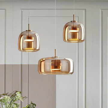 Three Retro Chandelier pendant lights with translucent amber glass shades and adjustable wires hang stylishly at varying heights against a white paneled wall, while greenery in the lower left corner adds a hint of nature to the contemporary scene.