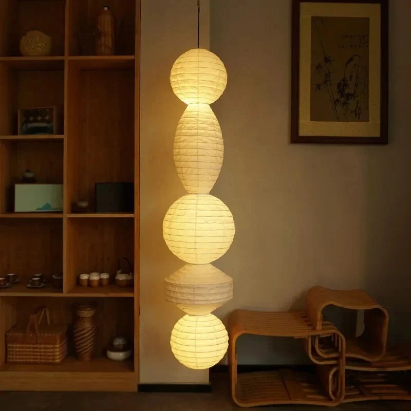 A cozy home interior showcases a Paper-Pasted Rice Paper Chandelier with LED bulbs and adjustable suspension. Nearby wooden shelves display vases, a wicker chair sits on the floor, and framed wall art completes the decor elegantly.