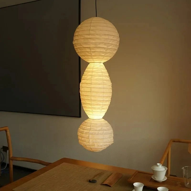 A cozy home interior showcases a Paper-Pasted Rice Paper Chandelier with LED bulbs and adjustable suspension. Nearby wooden shelves display vases, a wicker chair sits on the floor, and framed wall art completes the decor elegantly.