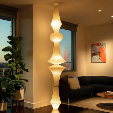 A modern living room showcases a Paper-Pasted Rice Paper Chandelier with adjustable suspension emitting a warm glow. Nearby, a large potted plant enhances the black L-shaped sofa and abstract art. Large windows reveal a cityscape at dusk.