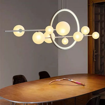 The Scandinavian Dimming Pendant Lamp, with round glowing bulbs and a circular LED frame on an adjustable rod, hangs above an oval wooden table. A decorative multicolored object adorns the table, surrounded by dark chairs, with a partially visible wooden door in the background.