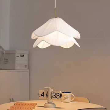 The Serenity Chandelier, with a petal shape and adjustable cable length, hangs over a round table adorned with a candle holder and mugs. The room is minimally decorated, featuring just a cabinet and boxes in the background.
