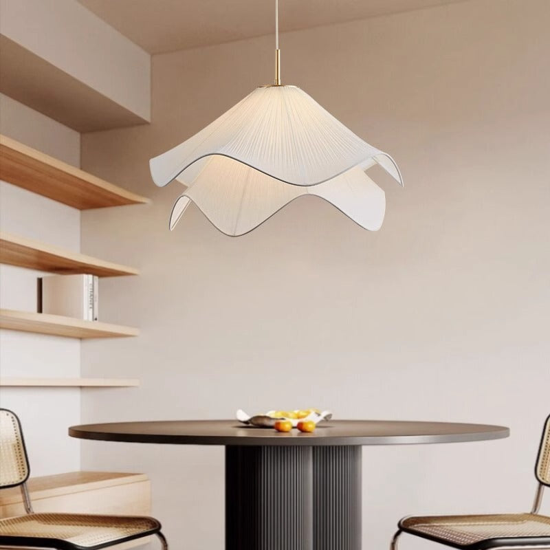 The Serenity Chandelier, with a petal shape and adjustable cable length, hangs over a round table adorned with a candle holder and mugs. The room is minimally decorated, featuring just a cabinet and boxes in the background.