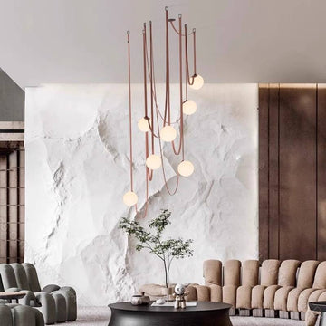 The modern living room showcases a large white textured wall and an elegant Branch Chandelier offering customizable dimming options. Plush, muted-tone seating encircles a round black coffee table next to a small tree, creating minimalist decor that emphasizes energy-efficient lighting.