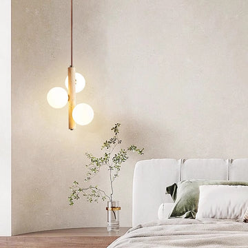 A minimalist bedroom showcases the Wabi Sabi Bubble Chandelier, a beige accent wall, and a cozy bed with white and green pillows. A small round table holds a clear vase with a leafy branch, enhancing the serene, stylish atmosphere.