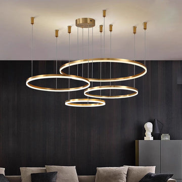 Modern chandelier with multiple circular LED rings hanging from the ceiling in a dark room with a gray sectional sofa and decorative elements on shelves.