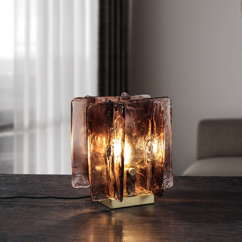 A Vintage brass glass table lamp with a hand-blown amber shade sits on a dark wooden table, emitting a warm, energy-efficient glow. Its positioned near a window with sheer curtains and has a beige couch in the background.