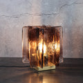 The Vintage brass glass table lamp, featuring a hand-blown amber glass shade and base, emits a warm glow as it stands on a dark wooden surface against a textured gray wall.
