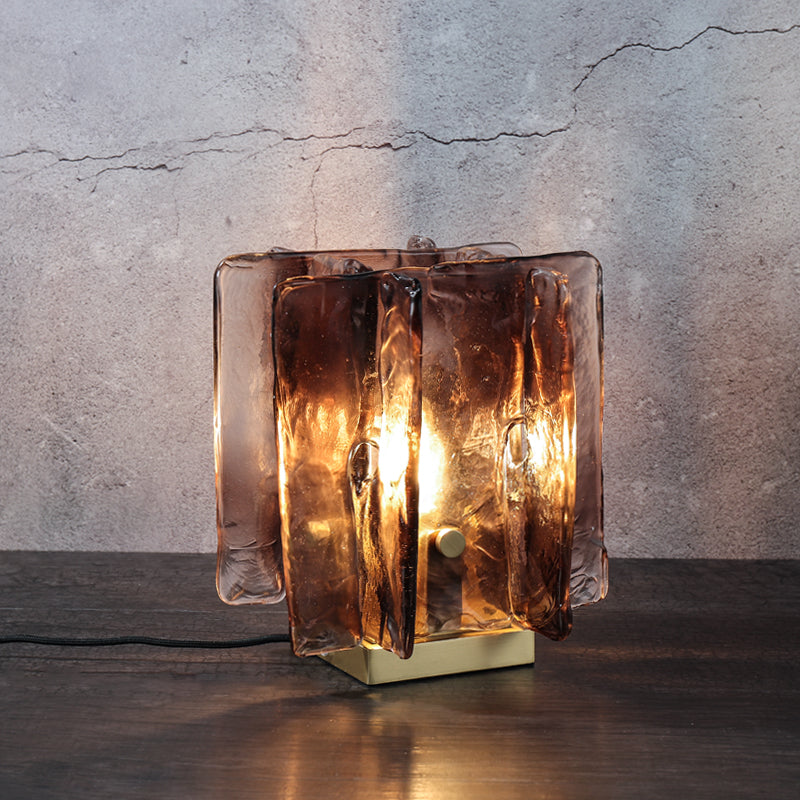 The Vintage brass glass table lamp, featuring a hand-blown amber glass shade and base, emits a warm glow as it stands on a dark wooden surface against a textured gray wall.