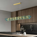 The Chandelier Series adds elegance to the modern kitchen with wooden cabinets and a black marble backsplash. This striking glass pendant light above the dark island features adjustable height and geometric blue and gold accents, complementing the white flower vase and glasses.