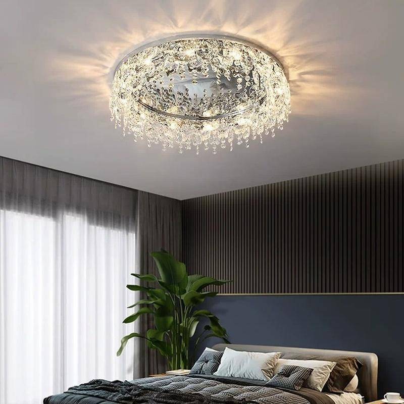 In an elegant bedroom, a Butterfly Crystal Ceiling Lamp with an LED bulb gently illuminates textured dark walls. A neatly made bed with plush pillows is below, while a tall plant on the left complements sheer curtains over the window.