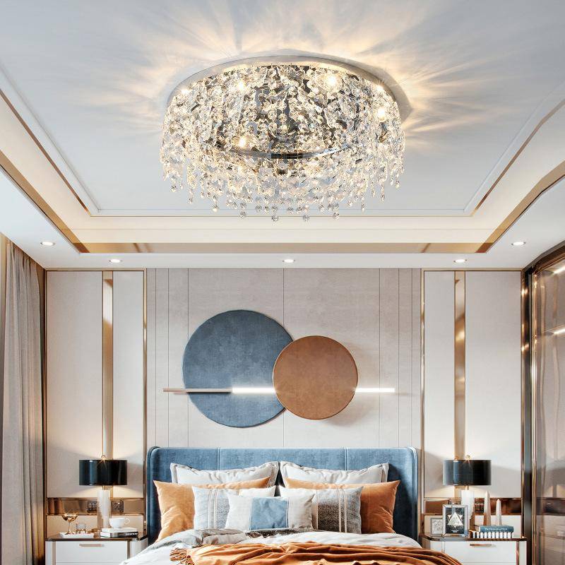 A luxurious bedroom features the Butterfly Crystal Ceiling Lamp, with blue and orange wall art above a plush bed adorned with decorative pillows, mirrored accents, sleek bedside lamps with LED bulbs, and neutral curtains.