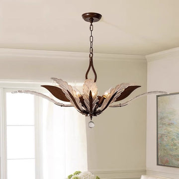 A chandelier with leaf-like metal and crystal elements hangs from a chain in a room with light-colored walls and a window in the background.
