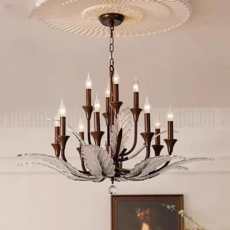 A chandelier with leaf-like metal and crystal elements hangs from a chain in a room with light-colored walls and a window in the background.