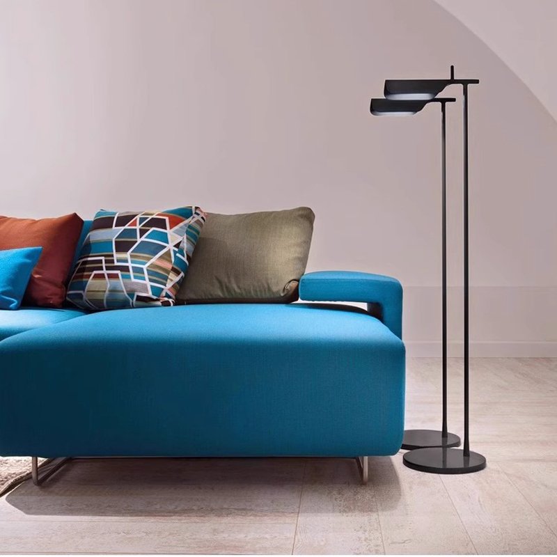 A modern living room includes a bright blue sofa adorned with colorful geometric-patterned cushions, complemented by a Simple floor lamp with a minimalist design standing on a light wooden floor. The walls, in an off-white shade, enhance the rooms tranquil ambiance.