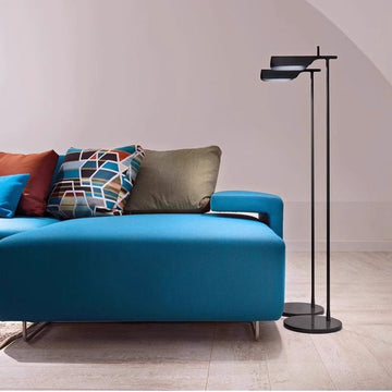A modern living room includes a bright blue sofa adorned with colorful geometric-patterned cushions, complemented by a Simple floor lamp with a minimalist design standing on a light wooden floor. The walls, in an off-white shade, enhance the rooms tranquil ambiance.