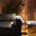 A cozy living room scene with a black leather sofa, draped with a gray blanket on the arm, is illuminated by a Simple floor lamp featuring a G9 LED bulb. The glass side table holds magazines, and a lush green plant complements the wood-paneled walls.