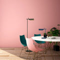 A modern room features a round white table with blue and pink chairs on a wooden floor. A Simple floor lamp casts a warm glow from its G9 LED bulb nearby, while a smaller desk lamp sits on the table. A green plant adds natures touch against the pink wall.