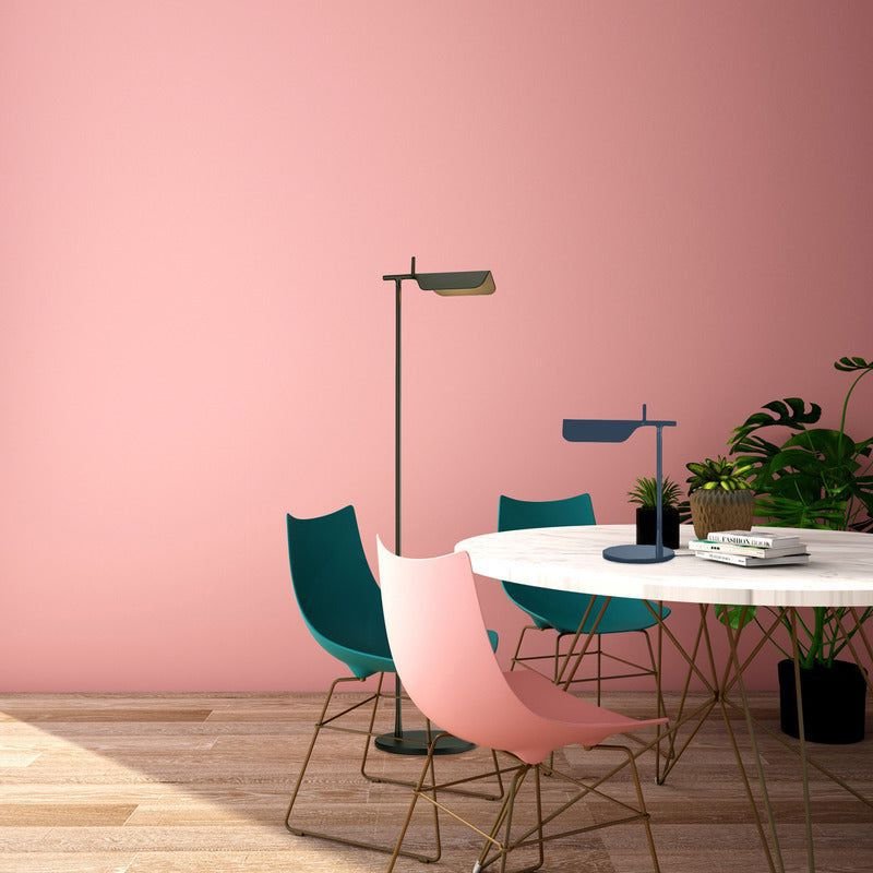 A modern room features a round white table with blue and pink chairs on a wooden floor. A Simple floor lamp casts a warm glow from its G9 LED bulb nearby, while a smaller desk lamp sits on the table. A green plant adds natures touch against the pink wall.