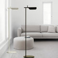 A minimalist living room features a light gray sofa and a circular white ottoman on a textured rug. Two Simple floor lamps with G9 LED bulbs stand near large windows with blinds, allowing diffused sunlight into the room.
