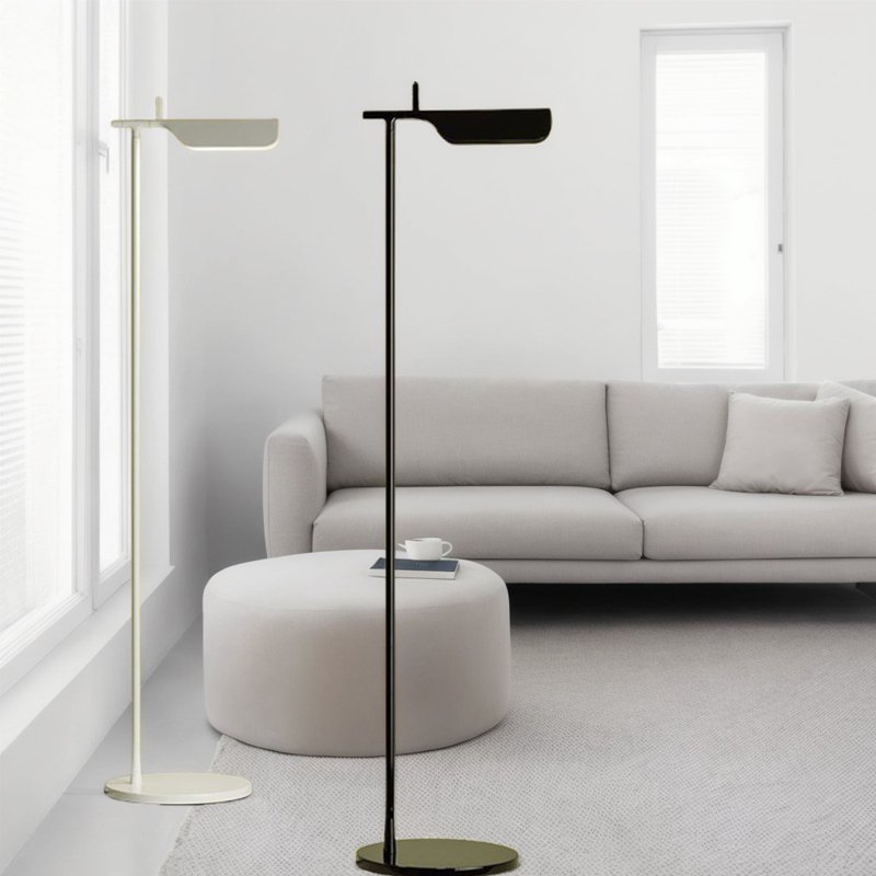 A minimalist living room features a light gray sofa and a circular white ottoman on a textured rug. Two Simple floor lamps with G9 LED bulbs stand near large windows with blinds, allowing diffused sunlight into the room.