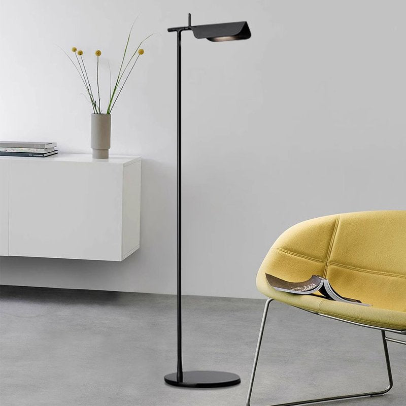 A modern living room boasts minimalist design with a Simple floor lamp using a G9 LED bulb, complemented by a white cabinet adorned with decor and vases. A yellow chair holds a book, set against gray walls and flooring for an effortlessly chic aesthetic.