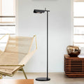 A modern, minimalist room features the Simple floor lamp with an angled shade and G9 LED bulb, illuminating the space. It stands beside a light-colored lounge chair with a gold frame, while a wooden cabinet holds a shiny apple-shaped decor piece. Large windows welcome natural light.