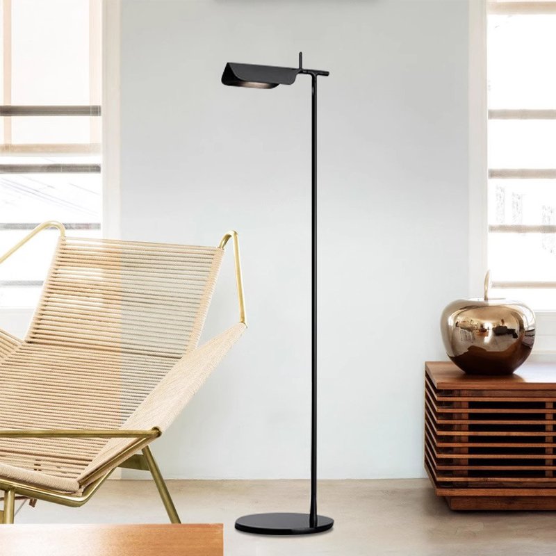 A modern, minimalist room features the Simple floor lamp with an angled shade and G9 LED bulb, illuminating the space. It stands beside a light-colored lounge chair with a gold frame, while a wooden cabinet holds a shiny apple-shaped decor piece. Large windows welcome natural light.