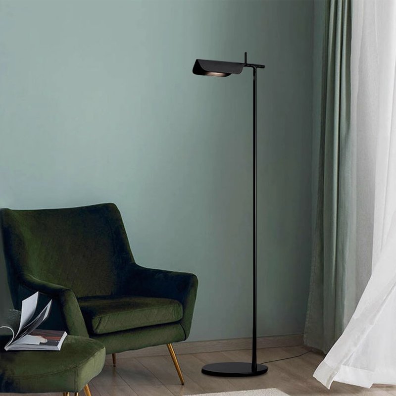 A modern living room showcases a dark green armchair and a Simple floor lamp with a G9 LED bulb for efficient lighting. A partially visible curtain adds texture, complemented by the light teal wall and wooden floor. A small table next to the chair holds several books, enhancing the minimalist design.
