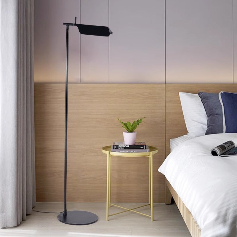A modern bedroom showcases a minimalist design with a sleek Simple floor lamp featuring a G9 LED bulb beside a small round table. The table hosts a potted plant and book. A neatly made bed with white and blue bedding sits adjacent to the light wood-paneled wall.