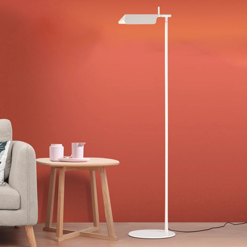 A Simple floor lamp with a minimalist design stands next to a small round wooden table holding a pink pitcher and cup. A beige sofa is partly visible on the left, while the warm orange background wall is softly illuminated by the G9 LED bulb.