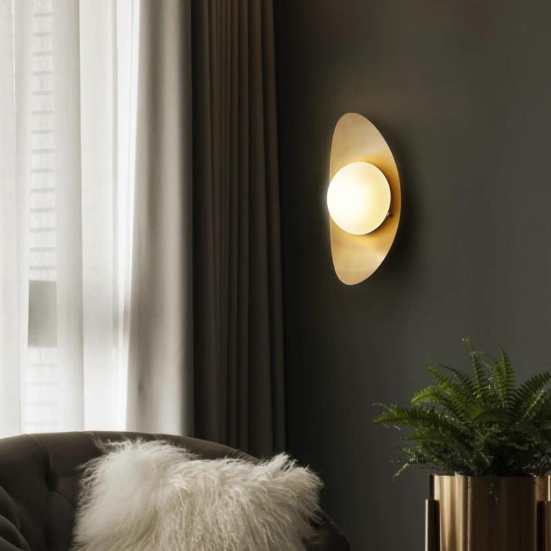 The elegant Metal Boat-Shaped Wall Light casts a warm glow on the dark wall, illuminating a nearby potted plant on a metallic stand. A plush chair with a fluffy white pillow is nestled beside sheer curtains that filter in soft daylight.